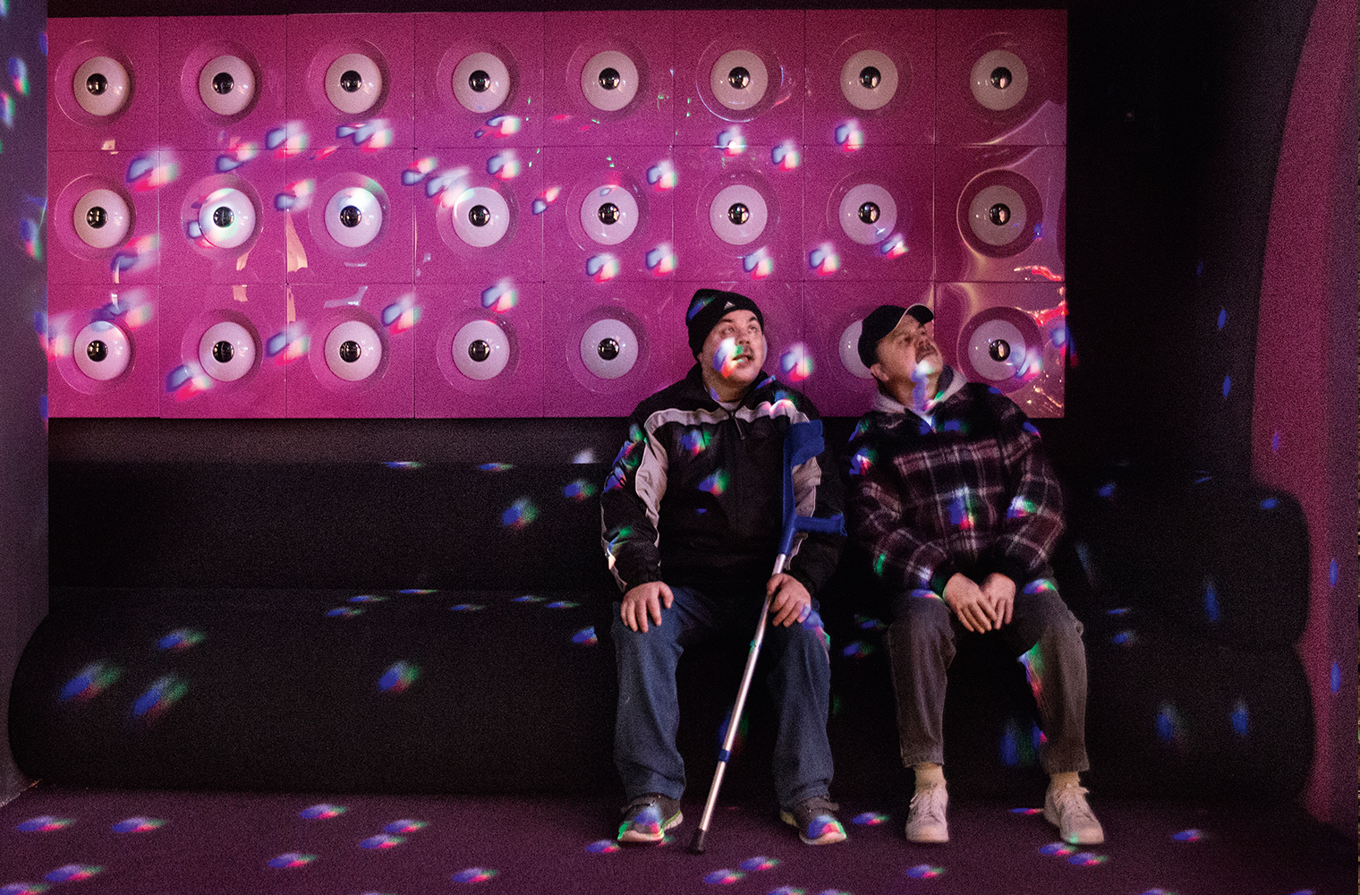 Die taubblinden Zwillingsbrüder Jörg und Rolf Fischer sitzen in einem in farbiges Licht getauchten Raum und spüren die Lichtreflexe. 