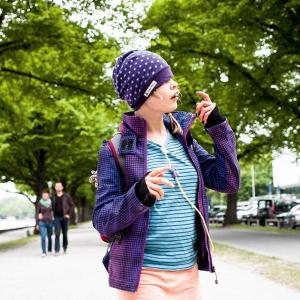 Ein Mädchen mit Haube, Jacke und über dem T-Shirt sichtbarem Ernährungsschlauch spaziert durch eine Straßenallee entlang eines Flusses. Im Hintergr...