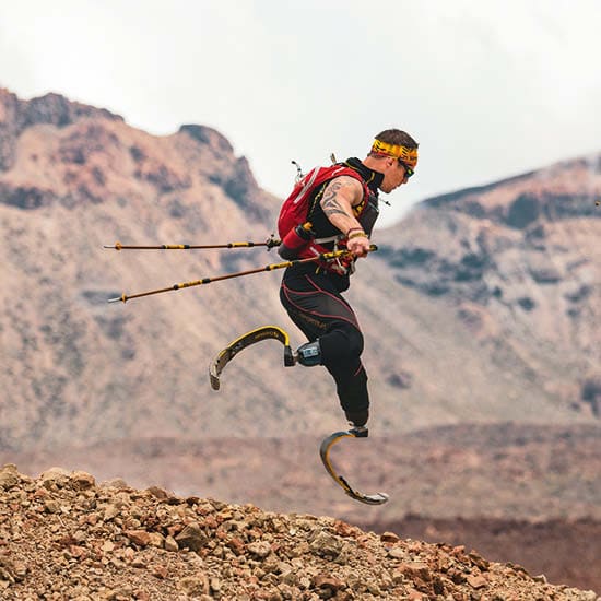 Ein Sportler mit Prothesen an beiden Beinen springt mit Wanderstöcken über steiniges Gelände. Er trägt eine rote Sportweste und eine Sonnenbrille....