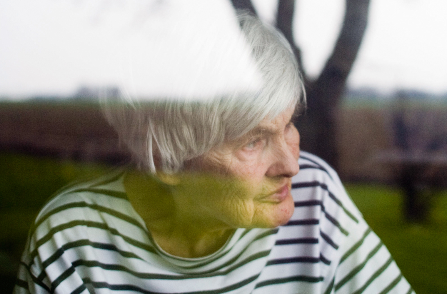 Eine ältere Dame steht am geschlossenen Fenster und schaut in den Garten.