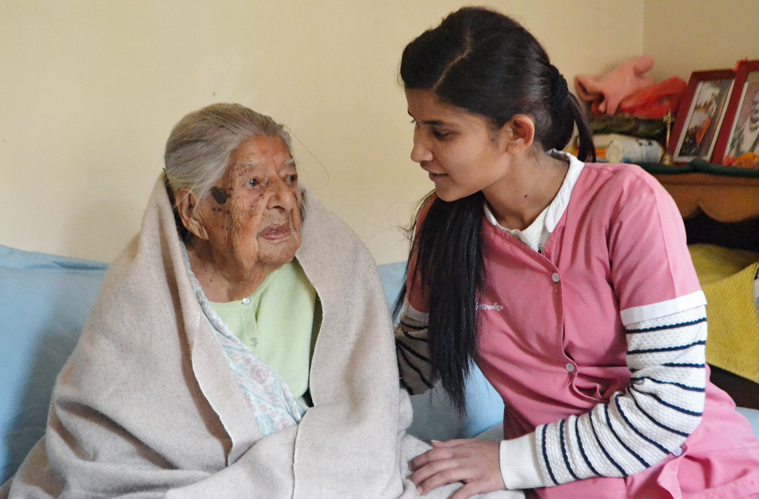 Lea Frei: Eine junge nepalesische Pflegerin sitzt neben einer älteren Frau, die in eine Decke eingehüllt ist, auf einer Couch und unterhält sich mi...